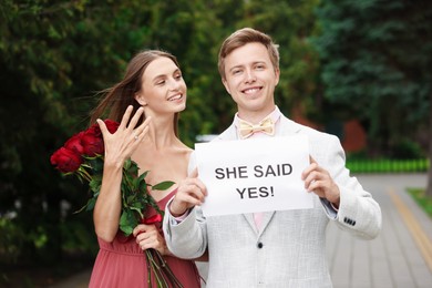 Photo of Smiling man holding sheet of paper with text She Said Yes and his happy beloved with bouquet of roses showing ring on engagement day outdoors