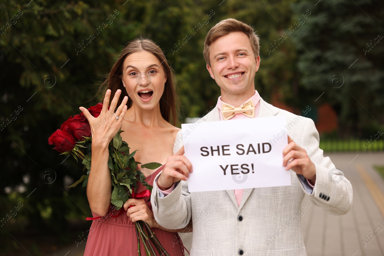 Smiling man holding sheet of paper with text She Said Yes and his emotional beloved with bouquet of roses showing ring on engagement day outdoors Photo of Smiling man holding sheet of paper with text She Said Yes and his emotional beloved with bouquet of roses showing ring on engagement day outdoors