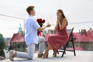 Photo of Man with engagement ring and bouquet of roses making marriage proposal to his happy girlfriend on balcony outdoors, low angle view