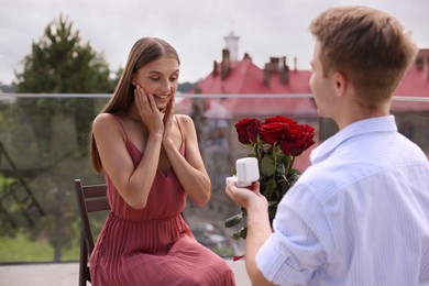 Photo of Man with engagement ring and bouquet of roses making marriage proposal to his happy girlfriend on balcony outdoors
