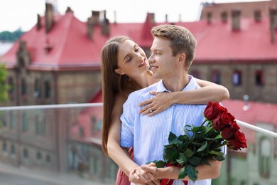 Photo of Making marriage proposal. Smiling woman with engagement ring holding bouquet of roses and her fiance hugging on balcony outdoors. Space for text