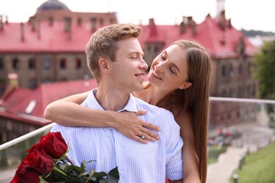 Photo of Making marriage proposal. Smiling woman with engagement ring holding bouquet of roses and her fiance hugging on balcony outdoors