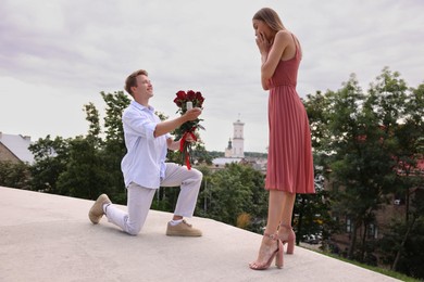 Photo of Smiling man with engagement ring and bouquet of roses making marriage proposal to his surprised girlfriend outdoors, low angle view