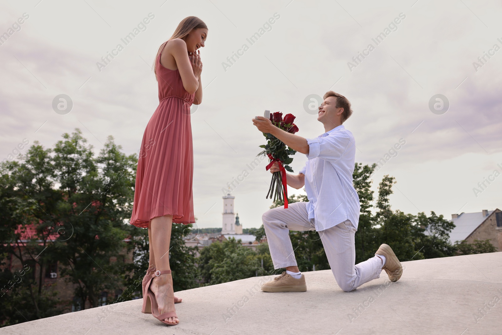 Smiling man with engagement ring and bouquet of roses making marriage proposal to his surprised girlfriend outdoors, low angle view Photo of Smiling man with engagement ring and bouquet of roses making marriage proposal to his surprised girlfriend outdoors, low angle view