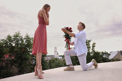 Photo of Smiling man with engagement ring and bouquet of roses making marriage proposal to his surprised girlfriend outdoors, low angle view
