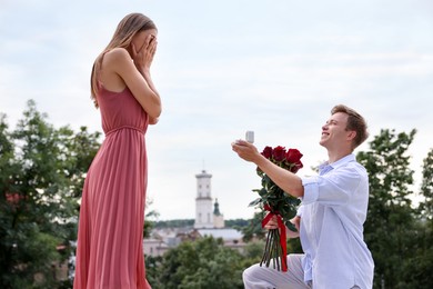 Photo of Smiling man with engagement ring and bouquet of roses making marriage proposal to his girlfriend outdoors, low angle view