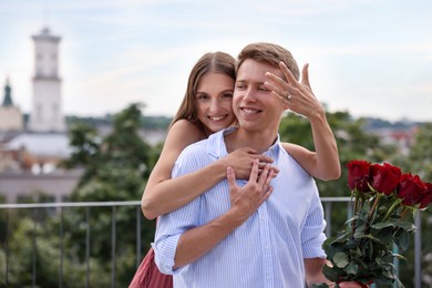 Photo of Marriage proposal. Smiling woman with engagement ring and her fiance holding bouquet of roses on balcony outdoors