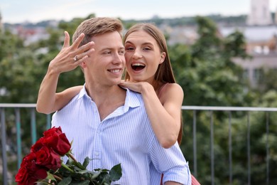 Photo of Marriage proposal. Excited woman with engagement ring and her fiance holding bouquet of roses on balcony outdoors