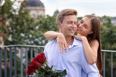 Photo of Marriage proposal. Smiling woman with engagement ring and her fiance holding bouquet of roses on balcony outdoors. Space for text