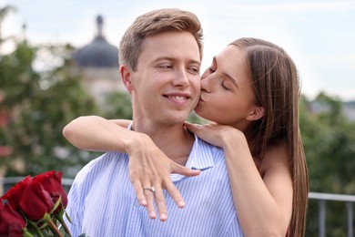 Photo of Marriage proposal. Woman with engagement ring kissing her fiance with bouquet of roses on balcony outdoors