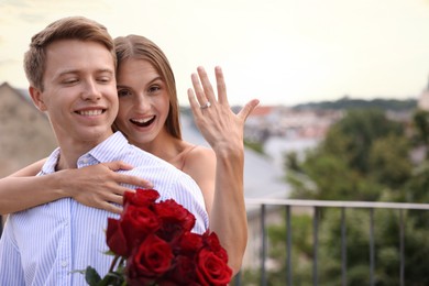 Photo of Marriage proposal. Happy woman showing engagement ring and her fiance holding bouquet of roses on balcony outdoors. Space for text