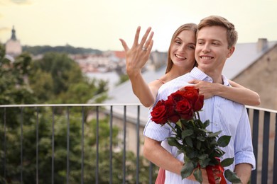 Photo of Marriage proposal. Smiling woman showing engagement ring and her fiance holding bouquet of roses on balcony outdoors. Space for text