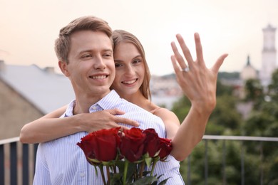 Photo of Marriage proposal. Smiling woman showing engagement ring and her fiance holding bouquet of roses on balcony outdoors, selective focus