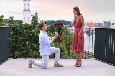 Photo of Man with engagement ring and bouquet of roses making marriage proposal to his happy girlfriend on balcony outdoors