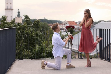 Photo of Man with engagement ring and bouquet of roses making marriage proposal to his surprised girlfriend on balcony outdoors. Space for text