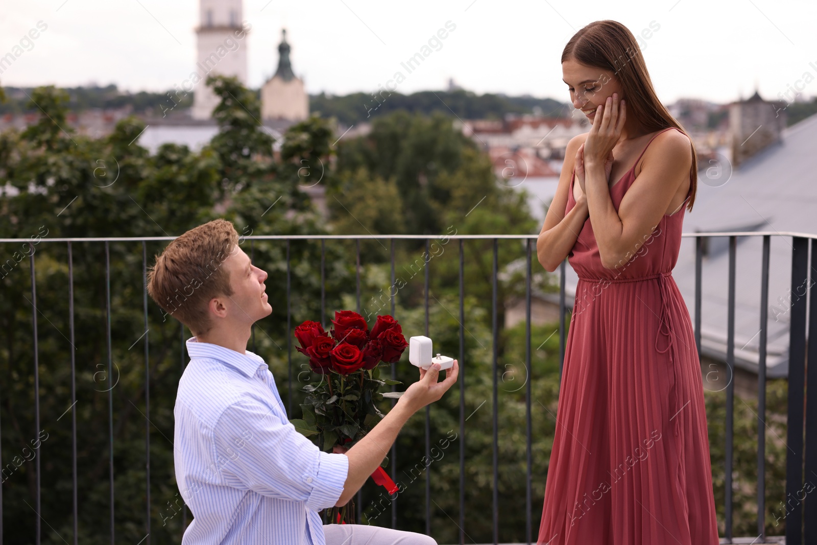 Man with engagement ring and bouquet of roses making marriage proposal to his smiling girlfriend on balcony outdoors Photo of Man with engagement ring and bouquet of roses making marriage proposal to his smiling girlfriend on balcony outdoors