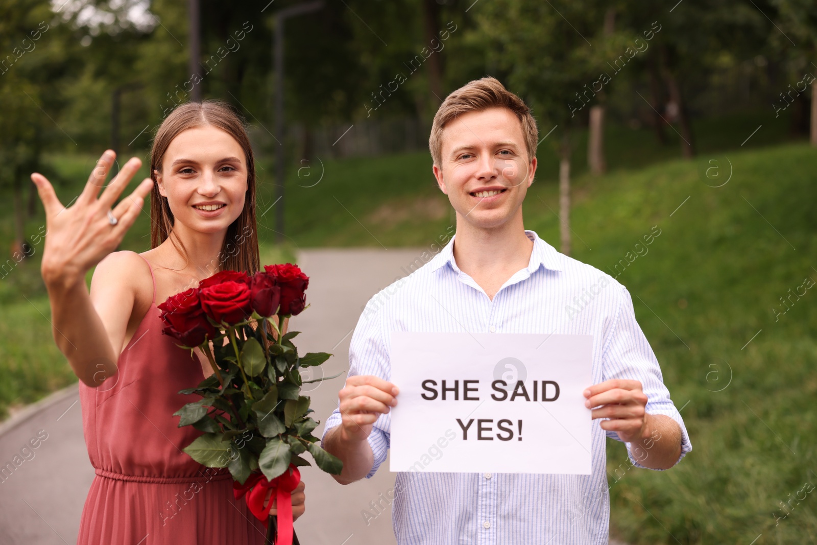 Smiling man holding sheet of paper with text She Said Yes and his happy beloved with bouquet of roses showing ring on engagement day outdoors Photo of Smiling man holding sheet of paper with text She Said Yes and his happy beloved with bouquet of roses showing ring on engagement day outdoors