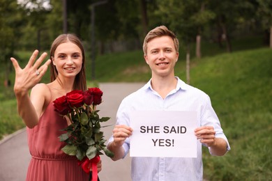 Photo of Smiling man holding sheet of paper with text She Said Yes and his happy beloved with bouquet of roses showing ring on engagement day outdoors