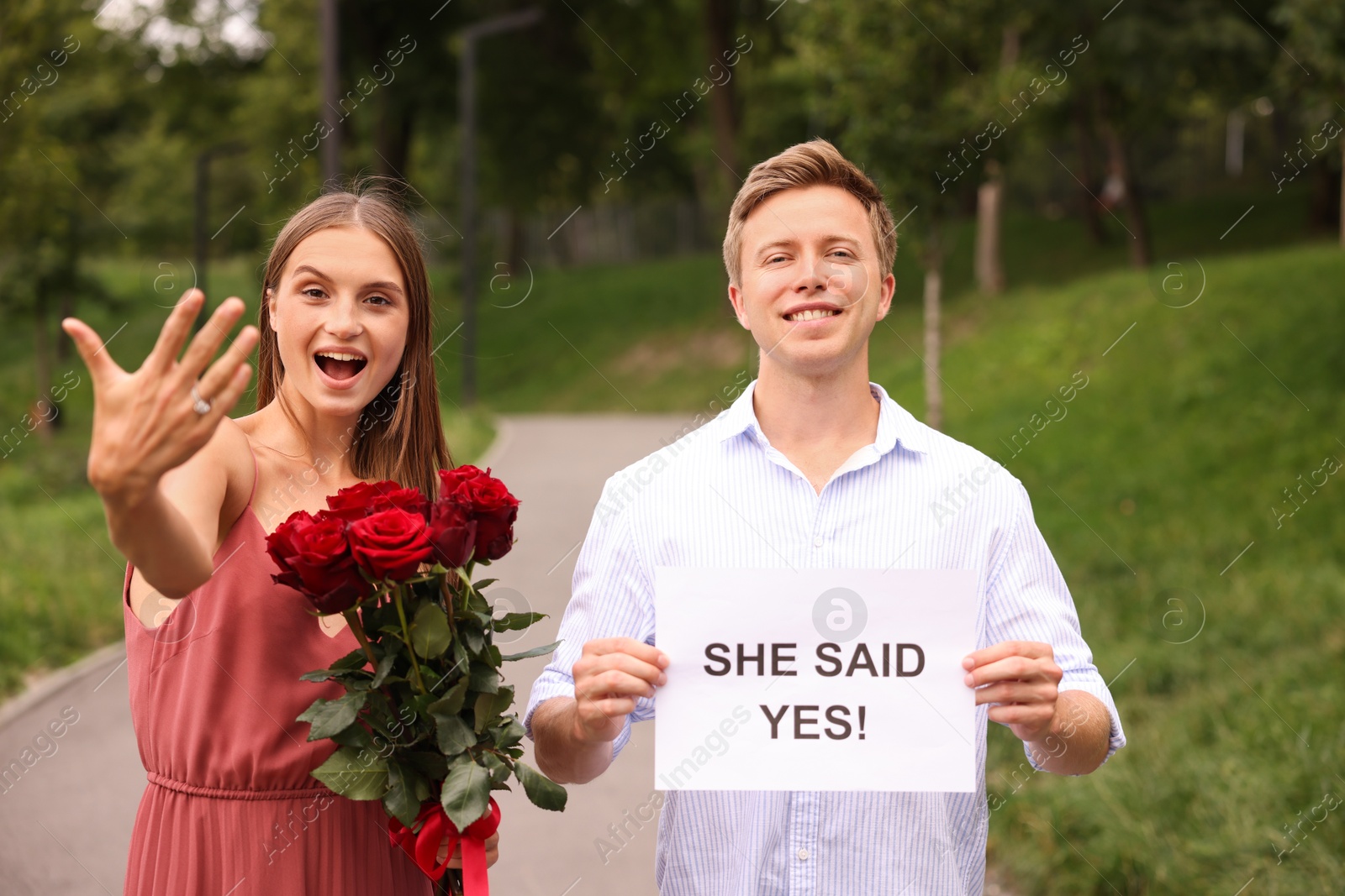 Smiling man holding sheet of paper with text She Said Yes and his excited beloved with bouquet of roses showing ring on engagement day outdoors Photo of Smiling man holding sheet of paper with text She Said Yes and his excited beloved with bouquet of roses showing ring on engagement day outdoors