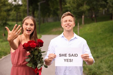 Photo of Smiling man holding sheet of paper with text She Said Yes and his excited beloved with bouquet of roses showing ring on engagement day outdoors