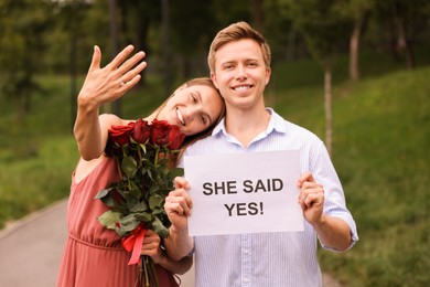 Photo of Smiling man holding sheet of paper with text She Said Yes and his happy beloved with bouquet of roses showing ring on engagement day outdoors