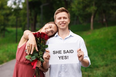 Photo of Smiling man holding sheet of paper with text She Said Yes and his excited beloved with bouquet of roses showing ring on engagement day outdoors