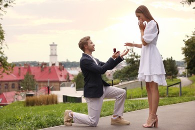 Photo of Smiling man with engagement ring making marriage proposal to his surprised girlfriend outdoors. Space for text