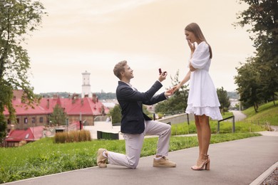 Photo of Smiling man with engagement ring making marriage proposal to his surprised girlfriend outdoors. Space for text