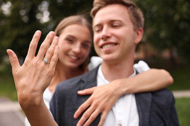 Photo of Marriage proposal. Smiling woman showing engagement ring and hugging her fiance outdoors, selective focus
