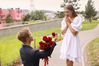 Photo of Man with engagement ring and bouquet of roses making marriage proposal to his surprised girlfriend outdoors
