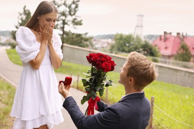 Photo of Man with engagement ring and bouquet of roses making marriage proposal to his surprised girlfriend outdoors