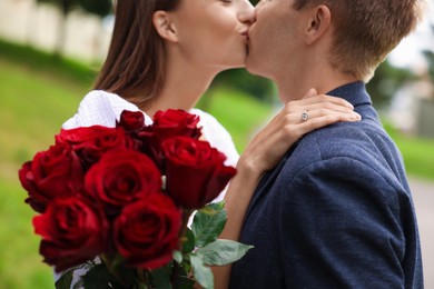 Photo of Marriage proposal. Man kissing his beloved with engagement ring and bouquet of roses outdoors, closeup