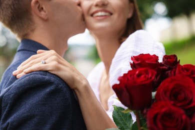 Photo of Marriage proposal. Man kissing his beloved with engagement ring and bouquet of roses outdoors, selective focus
