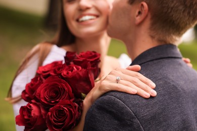 Photo of Marriage proposal. Man kissing his beloved with engagement ring and bouquet of roses outdoors, selective focus