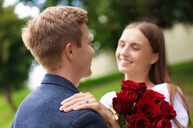 Photo of Marriage proposal. Smiling woman with engagement ring, bouquet of roses and her fiance outdoors, selective focus