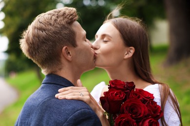 Photo of Marriage proposal. Man kissing his beloved with engagement ring and bouquet of roses outdoors