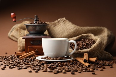 Photo of Cup of hot coffee, beans, grinder and cinnamon on brown background, closeup