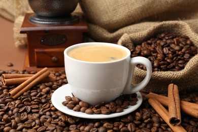 Photo of Cup of hot coffee, beans, grinder and cinnamon on brown background, closeup