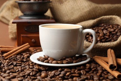 Photo of Cup of hot coffee, beans, grinder and cinnamon on brown background, closeup