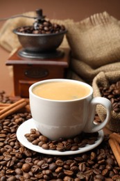 Photo of Cup of hot coffee, beans, grinder and cinnamon on brown background, closeup