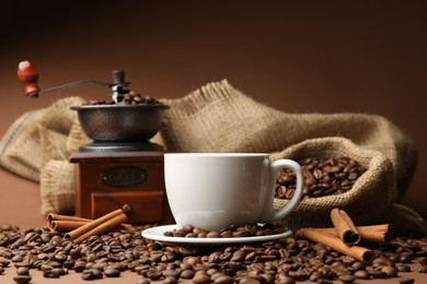 Photo of Cup of hot coffee, beans, grinder and cinnamon on brown background, closeup