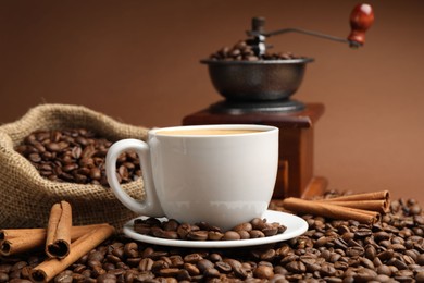 Photo of Cup of hot coffee, beans, grinder and cinnamon on brown background, closeup