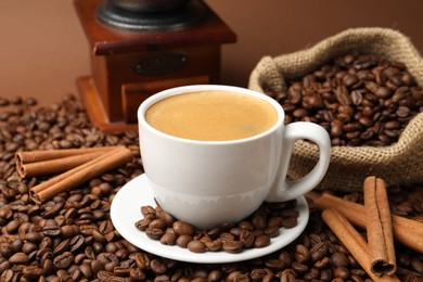 Photo of Cup of hot coffee, beans, grinder and cinnamon on brown background, closeup