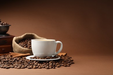 Photo of Cup of hot coffee, beans, grinder and cinnamon on brown background, space for text