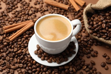 Photo of Cup of hot coffee, beans and cinnamon on brown background, closeup