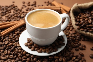 Photo of Cup of hot coffee, beans and cinnamon on brown background, closeup