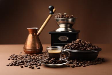 Photo of Cup of hot coffee, cezve, grinder and beans on brown background