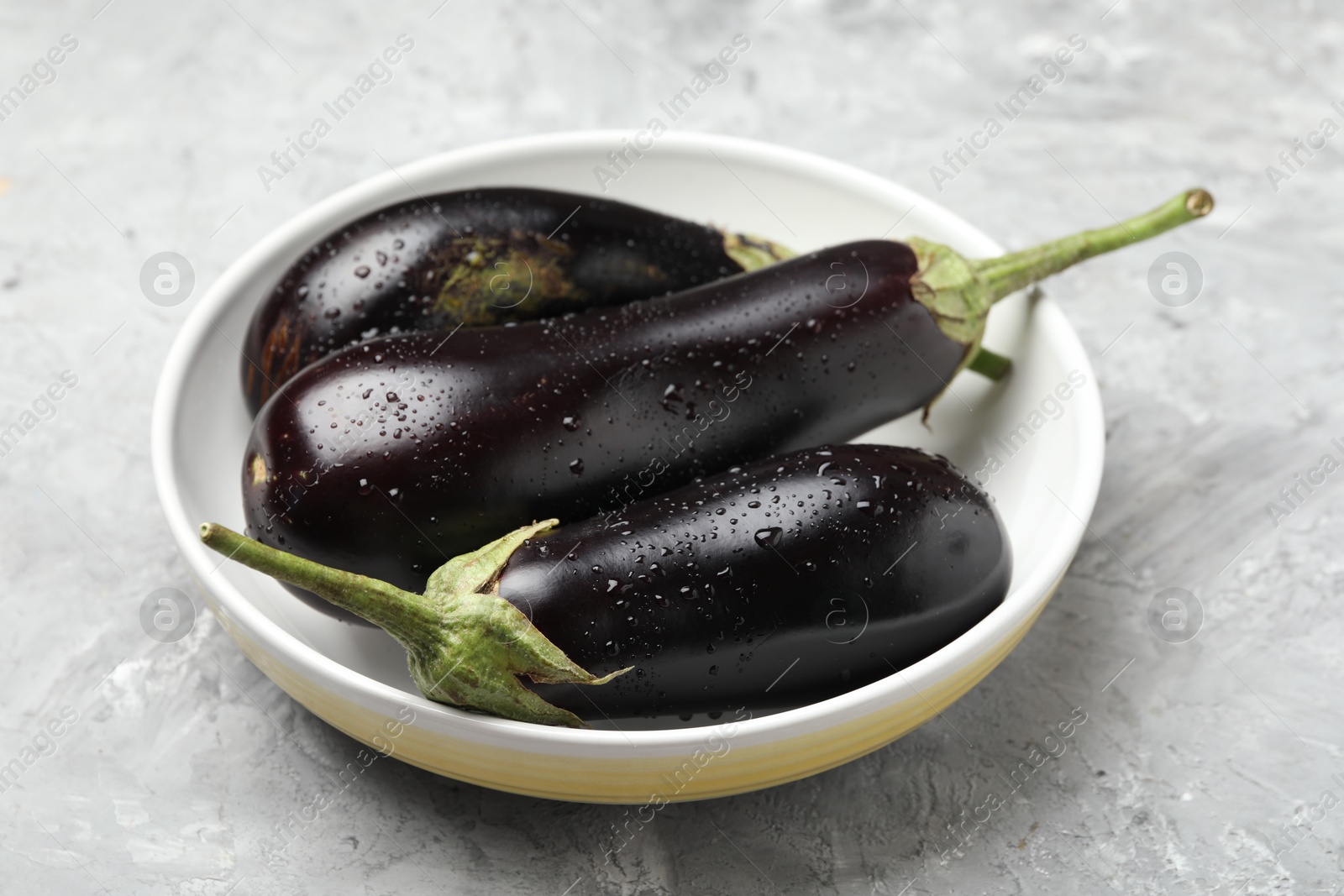 Fresh wet eggplants in bowl on grey textured table, closeup Photo of Fresh wet eggplants in bowl on grey textured table, closeup