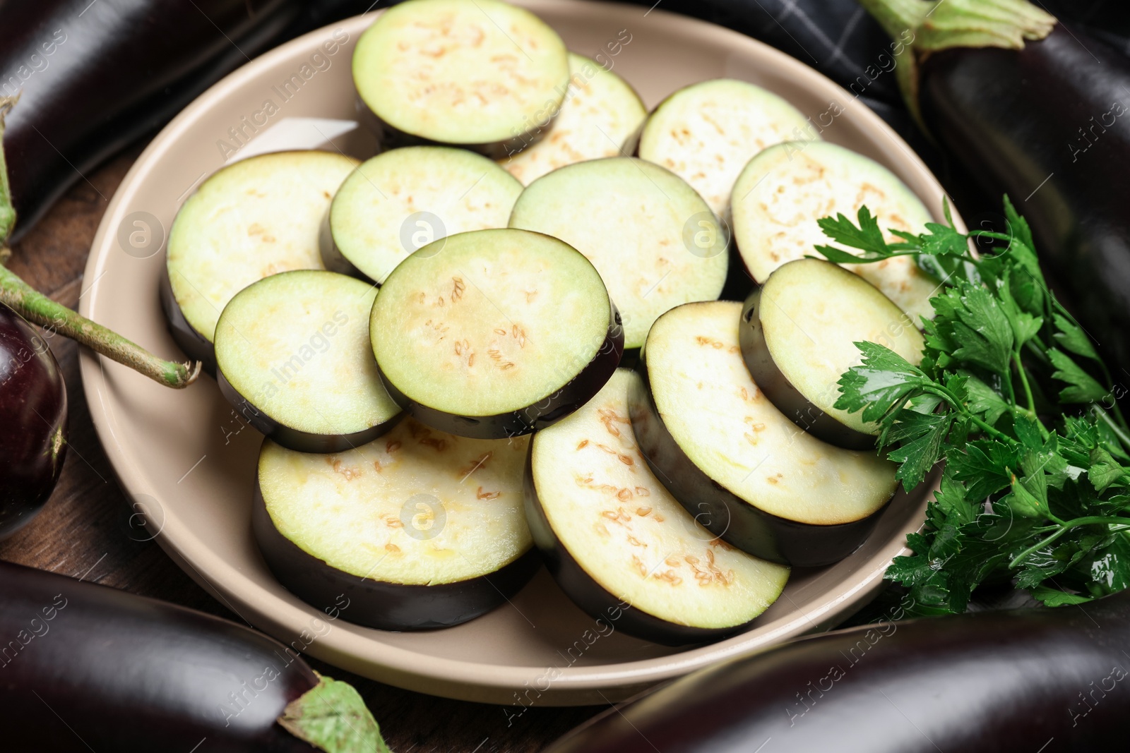 Photo of Slices of fresh eggplants surrounded by whole ones and parsley on wooden table, closeup