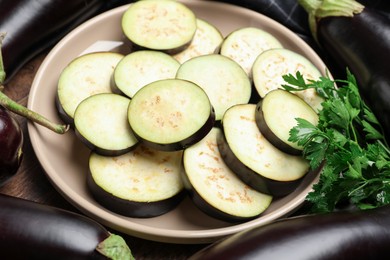 Photo of Slices of fresh eggplants surrounded by whole ones and parsley on wooden table, closeup